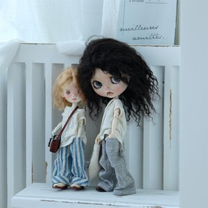 May include: Two dolls, one with long black curly hair and a white shirt and gray pants, and the other with short blonde hair and a white shirt and blue and white striped pants. The dolls are standing on a white shelf.