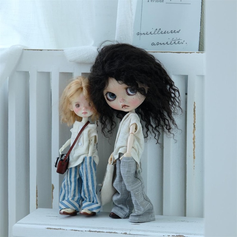 May include: Two dolls, one with long black curly hair and a white shirt and gray pants, and the other with short blonde hair and a white shirt and blue and white striped pants. The dolls are standing on a white shelf.