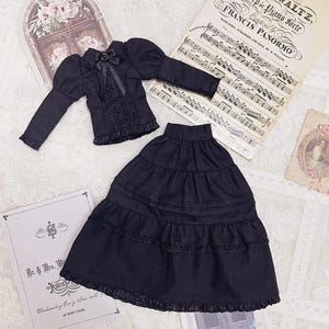 May include: A black doll outfit featuring a long-sleeved jacket with a bow and lace details, and a tiered skirt. The outfit is displayed with sheet music and a vintage-style card. The text on the card reads "Mr. & Mrs. W".
