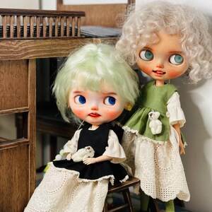 May include: Two custom art dolls with large eyes and unique hairstyles. One doll wears a black dress with a white lace skirt, the other a green dress with a white lace skirt. Both dolls have small stuffed animal accents.