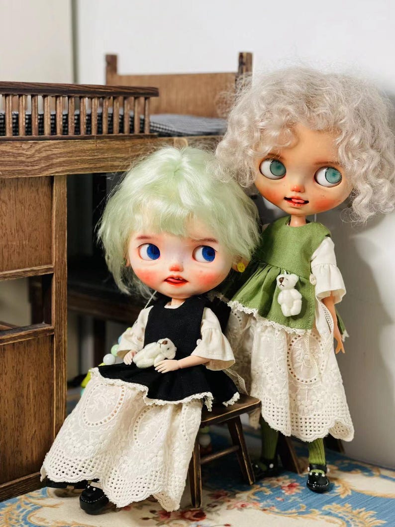May include: Two custom art dolls with large eyes and unique hairstyles. One doll wears a black dress with a white lace skirt, the other a green dress with a white lace skirt. Both dolls have small stuffed animal accents.