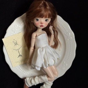 May include: A doll with auburn hair, wearing a white dress, leg warmers, and brown shoes, posed on a white plate. A small card with a sketch of a dress is next to the doll. The background is black.