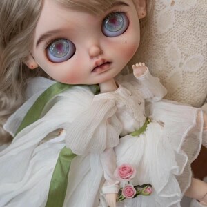 May include: A porcelain doll with blonde hair wearing a white dress with a green sash and holding a bouquet of pink roses.