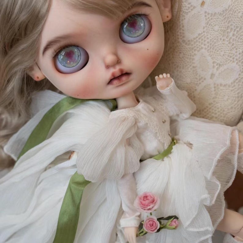 May include: A porcelain doll with blonde hair wearing a white dress with a green sash and holding a bouquet of pink roses.