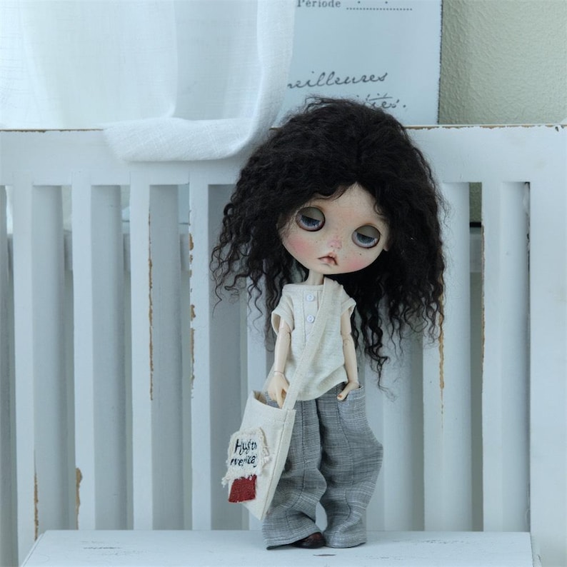 May include: A doll with long, curly black hair wearing a white shirt and gray pants. The doll is holding a small, white bag with the words "Auto de  merenda" embroidered on it.