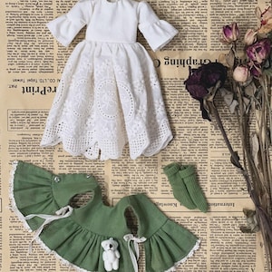 May include: A doll's dress set featuring a white eyelet dress with flared sleeves, a green cape with a white lace trim, and green socks. A small white teddy bear is attached to the cape. The items are arranged on a vintage newspaper background.