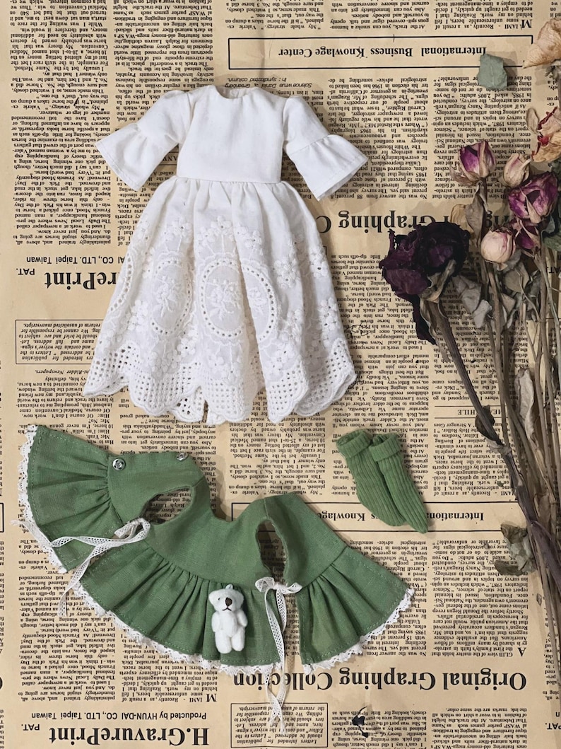 May include: A doll's dress set featuring a white eyelet dress with flared sleeves, a green cape with a white lace trim, and green socks. A small white teddy bear is attached to the cape. The items are arranged on a vintage newspaper background.