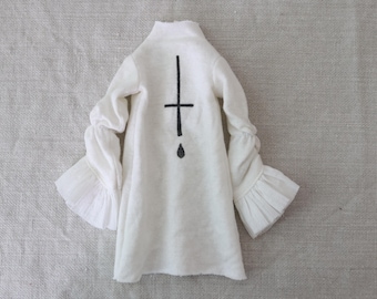 May include: A white long-sleeved garment with a high collar and flared sleeves. A black cross and teardrop design is on the back. The sleeves have a ruffled detail at the wrist.