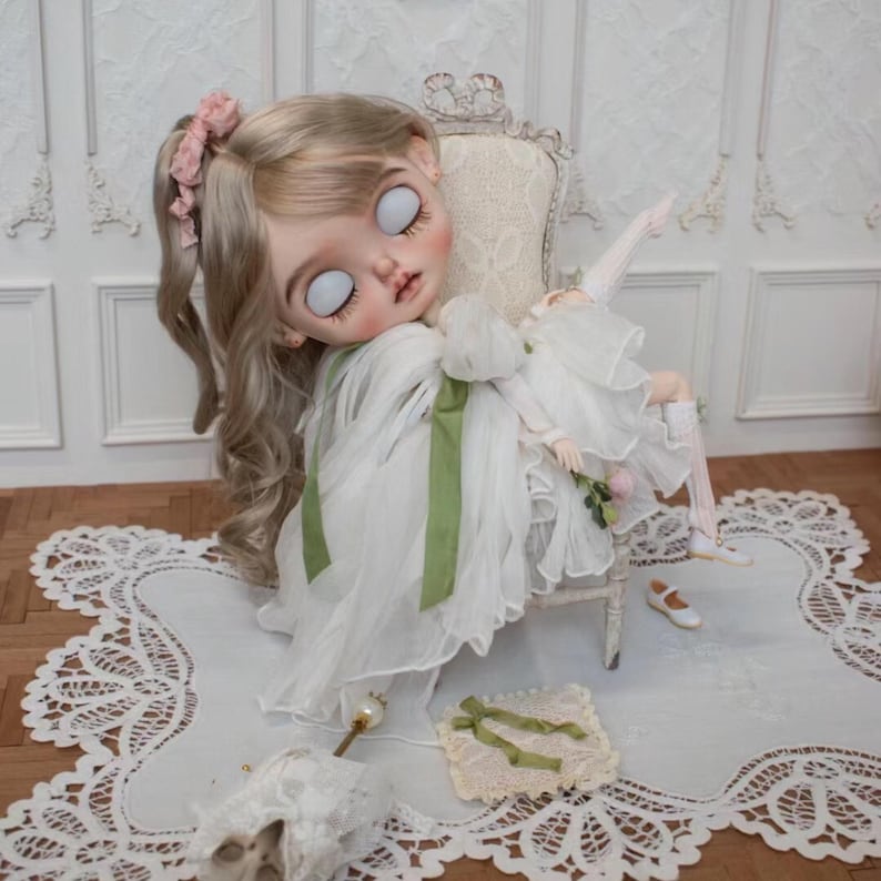 May include: A porcelain doll with blonde hair and a white dress sits on a white chair. The doll has large, white eyes and a green ribbon around her waist. The doll is sitting on a white lace doily.