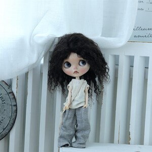 May include: A doll with long, curly, dark brown hair wearing a light brown sleeveless shirt and gray pants. The doll is sitting on a white chair with a white fabric draped over the back.
