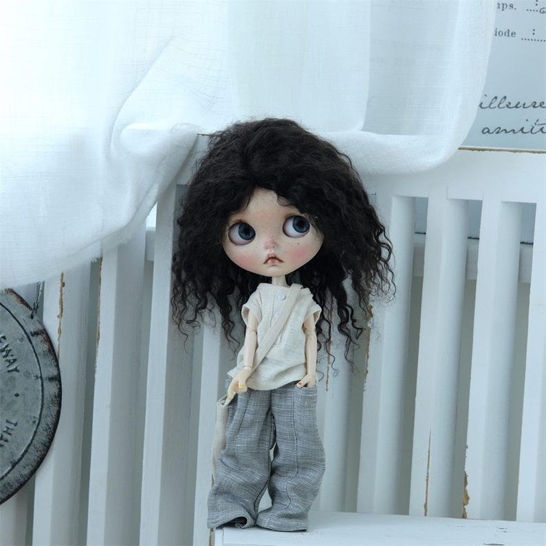 May include: A doll with long, curly, dark brown hair wearing a light brown sleeveless shirt and gray pants. The doll is sitting on a white chair with a white fabric draped over the back.