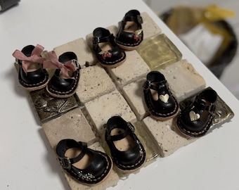 May include: A set of eight miniature black leather shoes with decorative details. Some shoes have pink bows, others have gold heart accents. The shoes are arranged on a tiled surface.