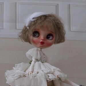 May include: A porcelain doll with blonde curly hair wearing a white dress with ruffles and a white hat with a feather. The doll is sitting on a brown box.