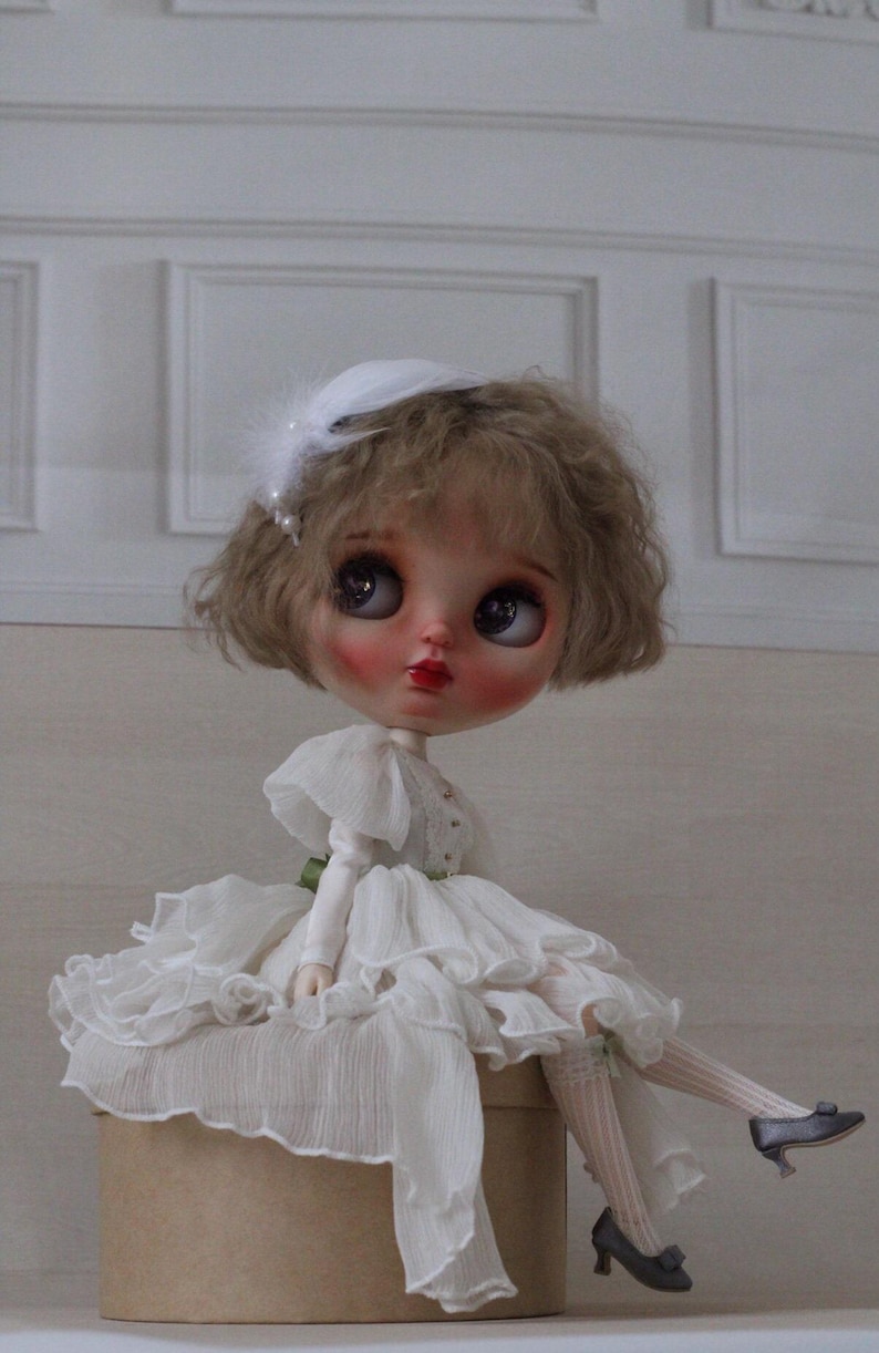 May include: A porcelain doll with blonde curly hair wearing a white dress with ruffles and a white hat with a feather. The doll is sitting on a brown box.