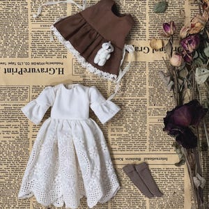 May include: A collection of doll clothing, including a white eyelet dress, a brown apron with a teddy bear, and brown socks. The items are arranged on a vintage newspaper background.