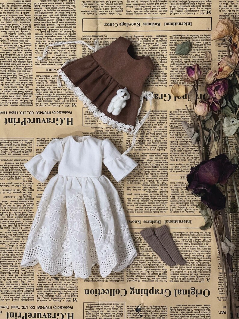 May include: A collection of doll clothing, including a white eyelet dress, a brown apron with a teddy bear, and brown socks. The items are arranged on a vintage newspaper background.
