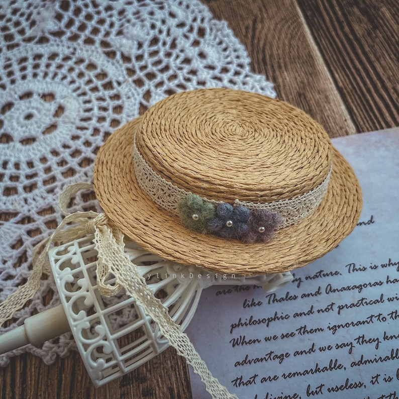 May include: A miniature straw hat with a white lace band and a cluster of colorful felt flowers. The hat is sitting on a white lace doily and a wooden surface.