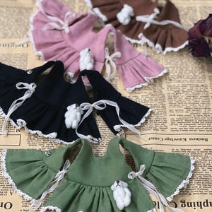 May include: A collection of doll dresses in various colors, including black, pink, brown, and green. Each dress features ruffled sleeves, white lace trim, and a small teddy bear embellishment. White ribbon ties add a delicate touch.