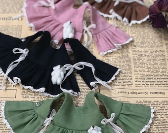 Op de afbeelding: Een verzameling poppenjurken in verschillende kleuren, waaronder zwart, roze, bruin en groen. Elke jurk heeft gerimpelde mouwen, witte kanten randen en een kleine teddybeerversiering. Witte linten voegen een delicate touch toe.