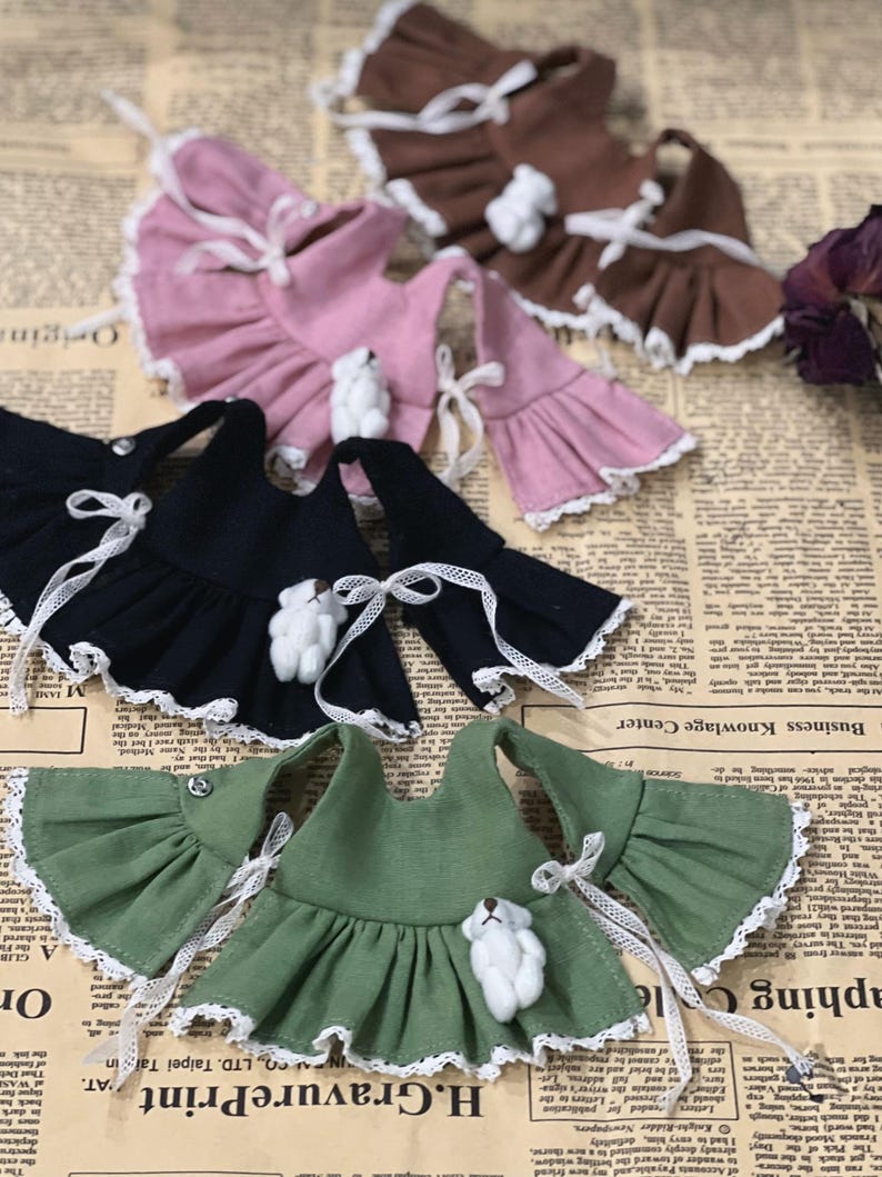 May include: A collection of doll dresses in various colors, including black, pink, brown, and green. Each dress features ruffled sleeves, white lace trim, and a small teddy bear embellishment. White ribbon ties add a delicate touch.