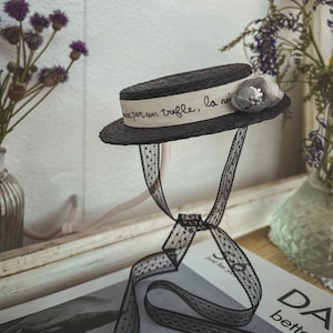 May include: A black straw hat with a white ribbon that says "prise par un truffle, la nuit" and a gray flower accent. The hat is tied with a black ribbon with white polka dots.