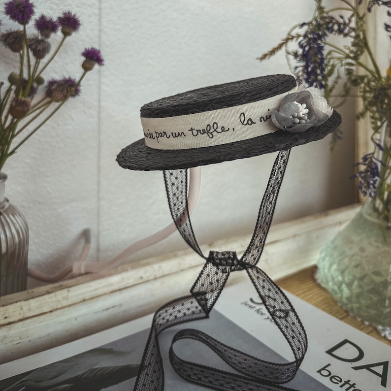 May include: A black straw hat with a white ribbon that says "prise par un truffle, la nuit" and a gray flower accent. The hat is tied with a black ribbon with white polka dots.