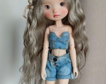 May include: A doll with long, wavy gray hair and a fair complexion. The doll wears a denim crop top and matching shorts. The doll's face has rosy cheeks and lips. The doll is wearing white socks.