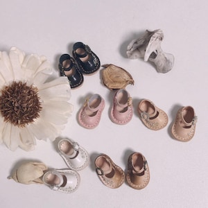 Op de afbeelding: Een assortiment miniatuur poppenschoenen in verschillende kleuren, waaronder zwart, roze, wit en bruin. De schoenen hebben delicate bloemendetails en gespen. De collectie bevat verschillende paren Mary Jane-schoenen.