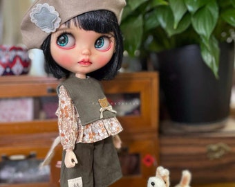 Puede incluir: Una muñeca con cabello negro y ojos azules que lleva un gorro marrón, una blusa con estampado floral, un chaleco verde y pantalones cortos verdes. La muñeca sostiene un pequeño perro de peluche.