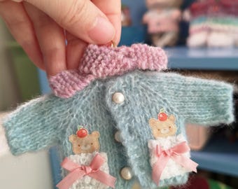 May include: A light blue knitted cardigan with a pink collar and two pockets. Each pocket has a white trim and a small embroidered bear with a red apple on its head. The cardigan has pearl buttons.