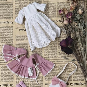 May include: A collection of doll clothes, including a white eyelet dress, a pink outfit with a teddy bear, a purse with a bow, and pink socks. The items are arranged on a vintage newspaper background.