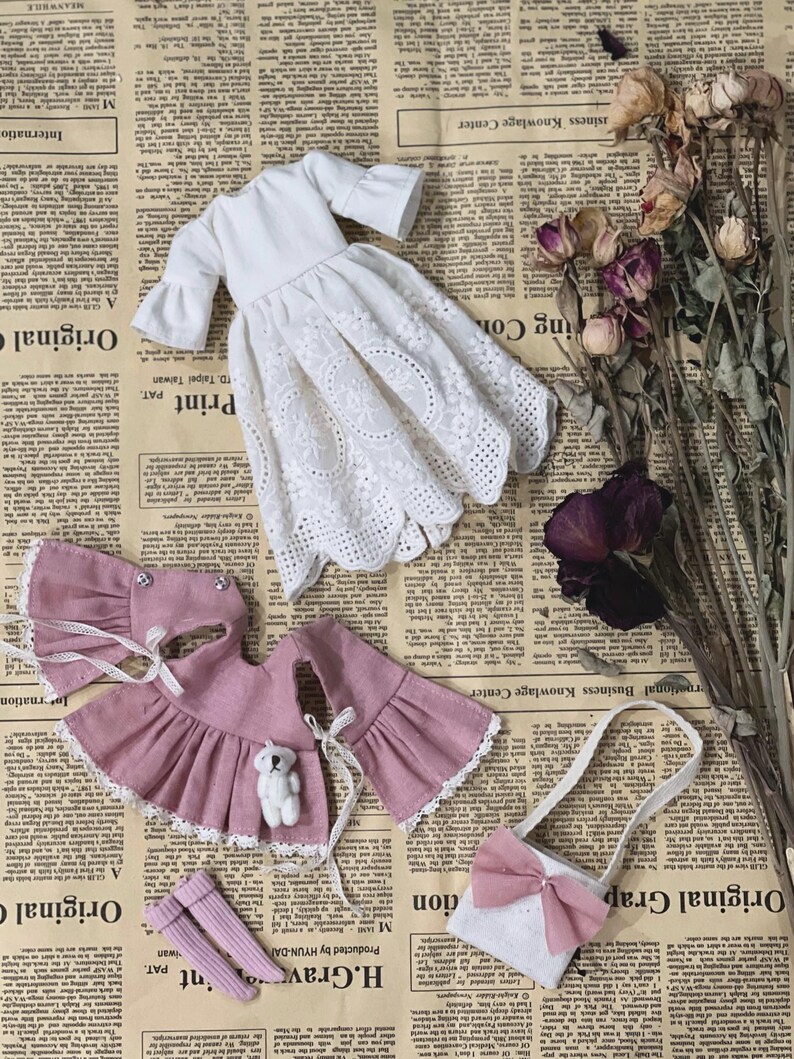 May include: A collection of doll clothes, including a white eyelet dress, a pink outfit with a teddy bear, a purse with a bow, and pink socks. The items are arranged on a vintage newspaper background.