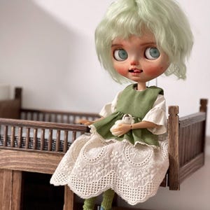 May include: A Blythe doll with mint green hair, wearing a white dress with lace detailing and a green vest. The doll is holding a small white stuffed animal and is seated on a miniature wooden bed frame. The doll is wearing teal shoes and green socks.