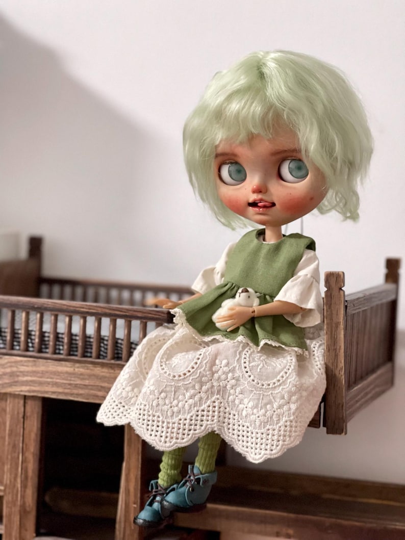 May include: A Blythe doll with mint green hair, wearing a white dress with lace detailing and a green vest. The doll is holding a small white stuffed animal and is seated on a miniature wooden bed frame. The doll is wearing teal shoes and green socks.