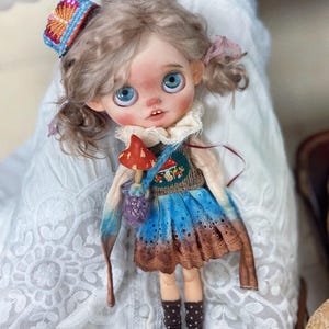 Op de afbeelding: Een handgemaakte pop met grote blauwe ogen, gekleed in een blauwe en bruine jurk, een gebreid vest en een kleurrijke hoofdband. De pop heeft een klein paddenstoelvormig accessoire en een paarse tas. Het haar van de pop is opgestoken in staartjes.