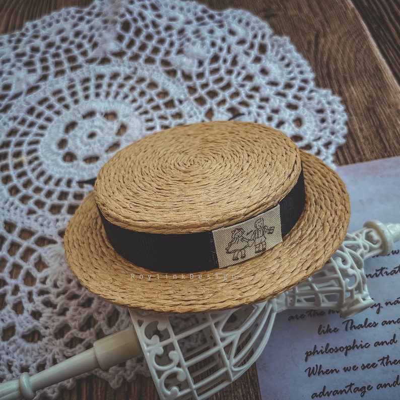 May include: A straw boater hat with a black band and a small embroidered design of two figures holding hands. The hat is sitting on a white crocheted doily.