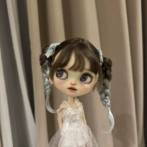 May include: A doll with brown hair styled in pigtails with blue accents. The doll has large eyes and is wearing a white dress with a sheer overlay. The doll's face has a delicate, painted appearance.