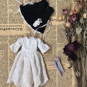 May include: A doll's outfit featuring a white dress with a lace skirt, a black vest with a teddy bear, and gray socks. The dress has flared sleeves. The items are arranged on a newspaper background.