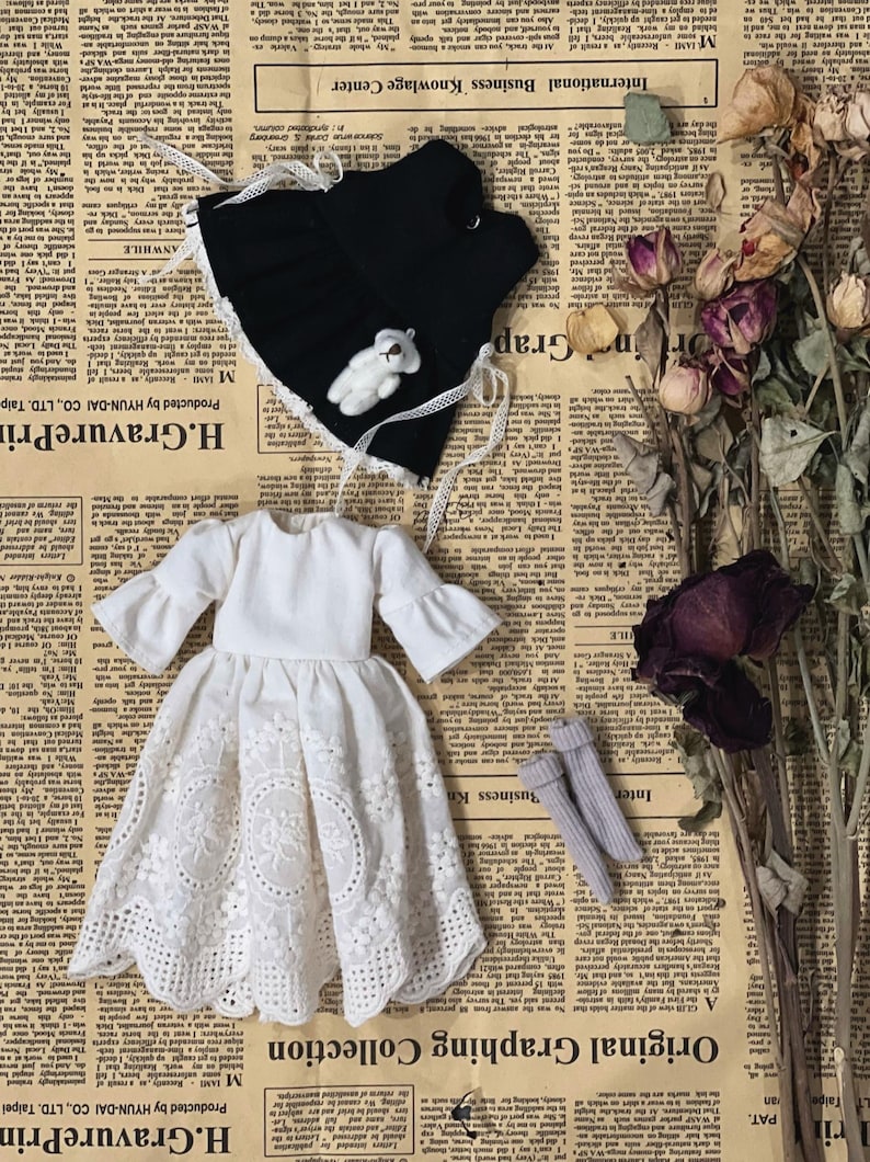 May include: A doll's outfit featuring a white dress with a lace skirt, a black vest with a teddy bear, and gray socks. The dress has flared sleeves. The items are arranged on a newspaper background.