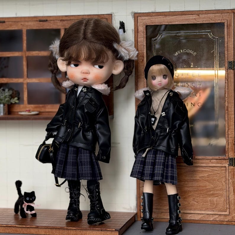 May include: Two dolls dressed in matching outfits stand side-by-side. Each doll wears a black faux leather jacket, a plaid skirt, and black boots. One doll carries a black purse. A black cat figurine with a pink bow is in the foreground.