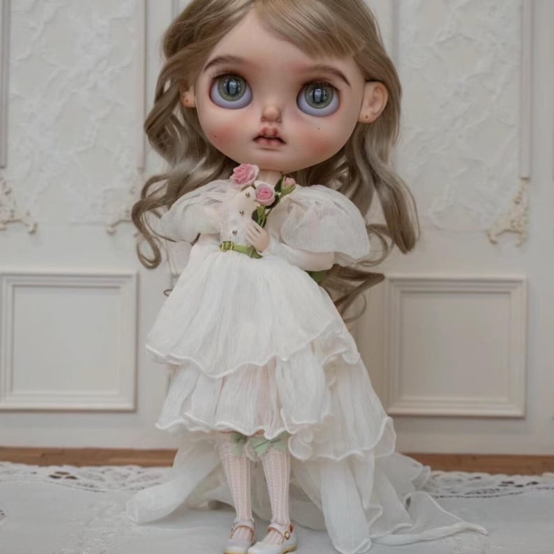 May include: A porcelain doll with blonde hair wearing a white dress with lace trim and holding a bouquet of pink flowers.