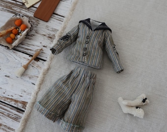 Puede incluir: Un conjunto para muñeco que consta de una camisa de manga larga abotonada con cuello marinero oscuro y pantalones a rayas a juego en tonos azul, beige y gris. El set incluye un par de calcetines de punto color crema con lazos negros.