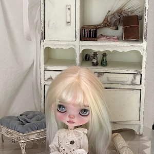 May include: A doll with long blonde hair and large blue eyes sits in front of a white cabinet with open shelves. The doll wears a white dress with lace details. The cabinet is decorated with miniature items, including a red watering can and small glass bottles.