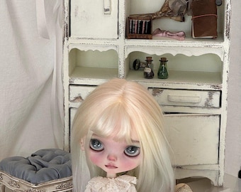 May include: A doll with long blonde hair and large blue eyes sits in front of a white cabinet with open shelves. The doll wears a white dress with lace details. The cabinet is decorated with miniature items, including a red watering can and small glass bottles.
