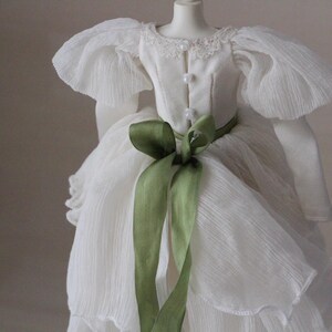 May include: A white dress with a green ribbon tied around the waist. The dress has a ruffled skirt and puffed sleeves.