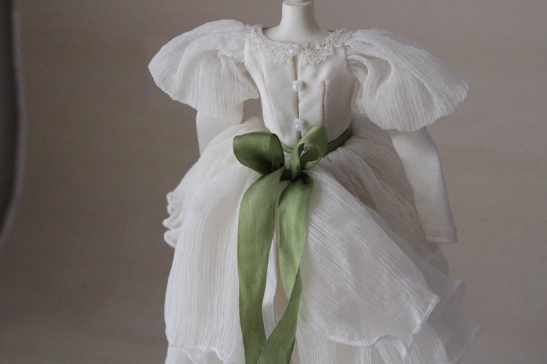 May include: A white dress with a green ribbon tied around the waist. The dress has a ruffled skirt and puffed sleeves.