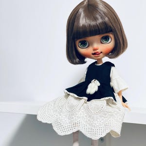 May include: A doll with brown hair and large green eyes, wearing a black dress with a white lace skirt and black shoes. The doll has a small white teddy bear on the dress. The doll is sitting on a white surface.
