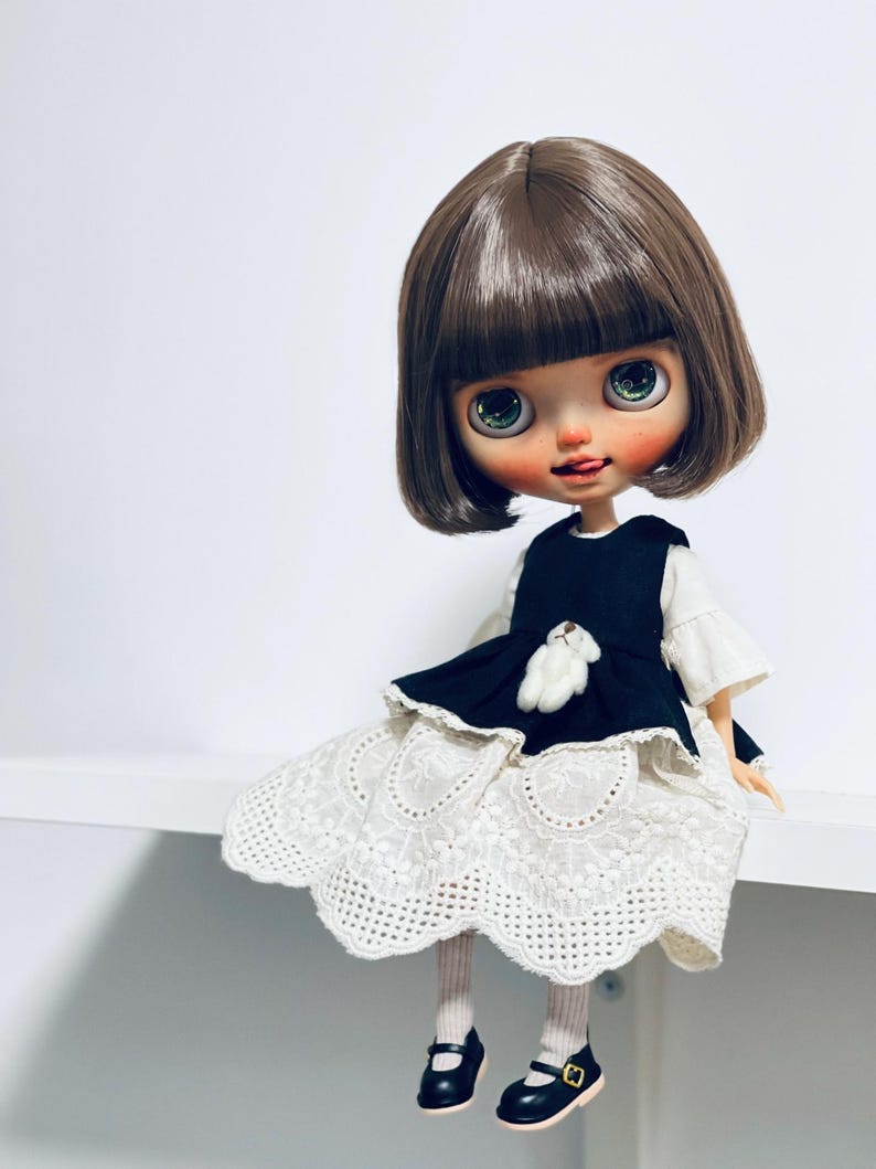 May include: A doll with brown hair and large green eyes, wearing a black dress with a white lace skirt and black shoes. The doll has a small white teddy bear on the dress. The doll is sitting on a white surface.