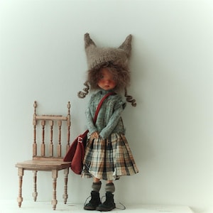 May include: A handmade doll dressed in a grey sweater, plaid skirt, and a brown hat with pointed ears. The doll has curly hair and carries a red bag. A small wooden chair is positioned nearby.