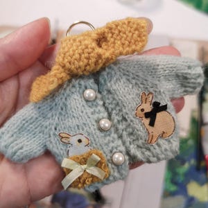 Puede incluir: Un suéter de punto azul claro con una bufanda amarilla y dos conejos bordados. El suéter tiene botones de perla y un lazo amarillo.